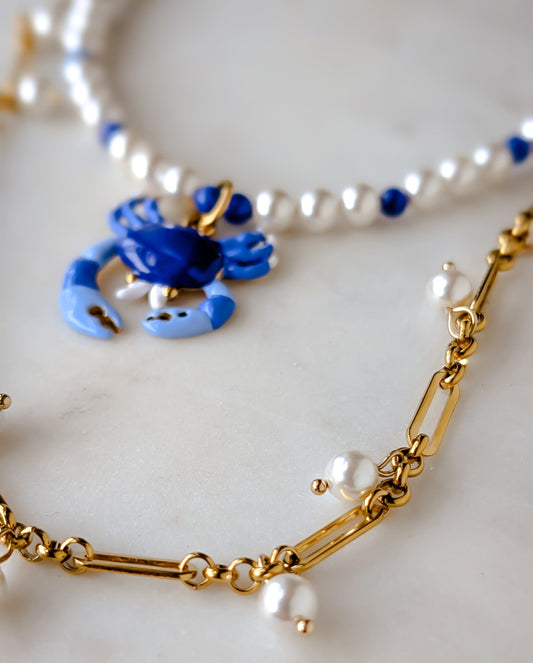 Gold chain necklace with blue crab pendant and pearl necklace on a white background