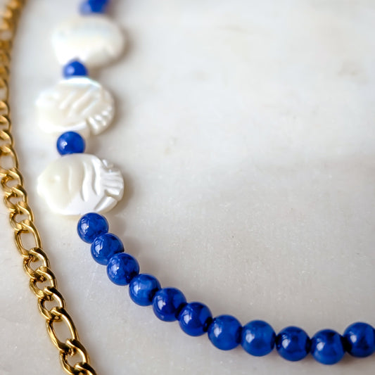 Close-up of a blue beaded bracelet with gold chain and white shell-like beads on a light background
