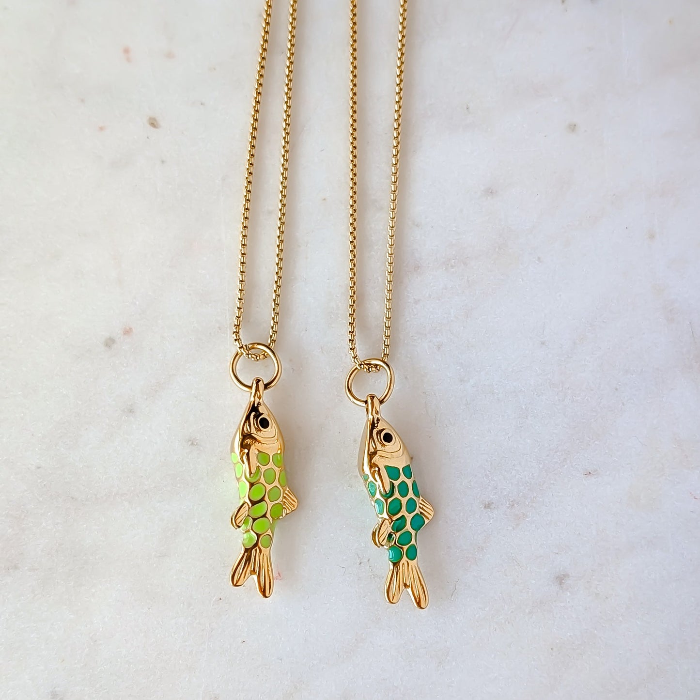 Two gold fish-shaped pendants Aqua and green enamel, and on chains against a light background