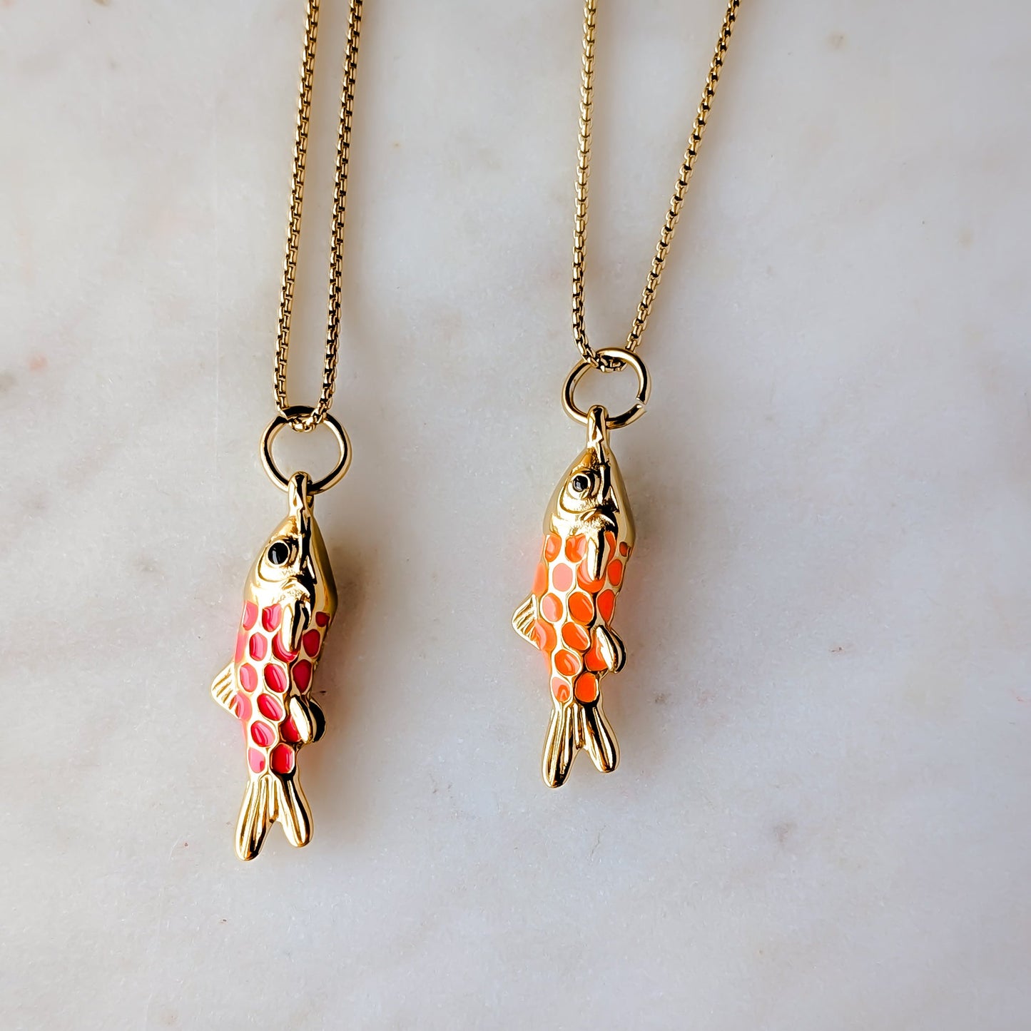 Two gold enamel orange and hot pink fish-shaped pendants on chains against a marble background