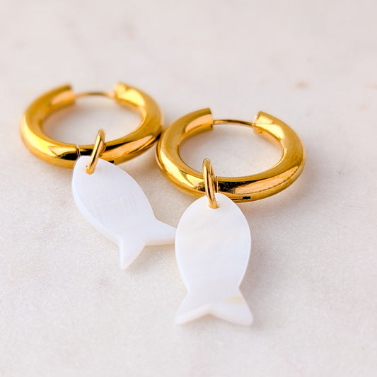 Gold hoop earrings with white fish charms on a light background